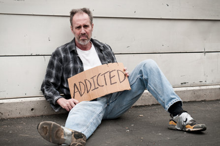A homeless man holding a sign that says Addicted.