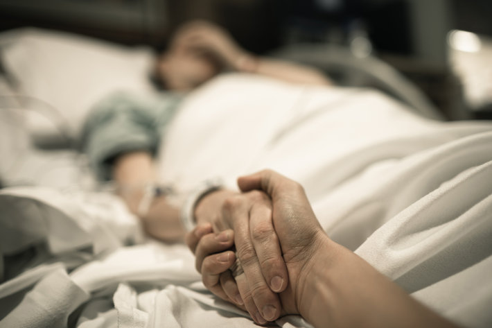 Sick woman lying in hospital bed with hand being held by love one.